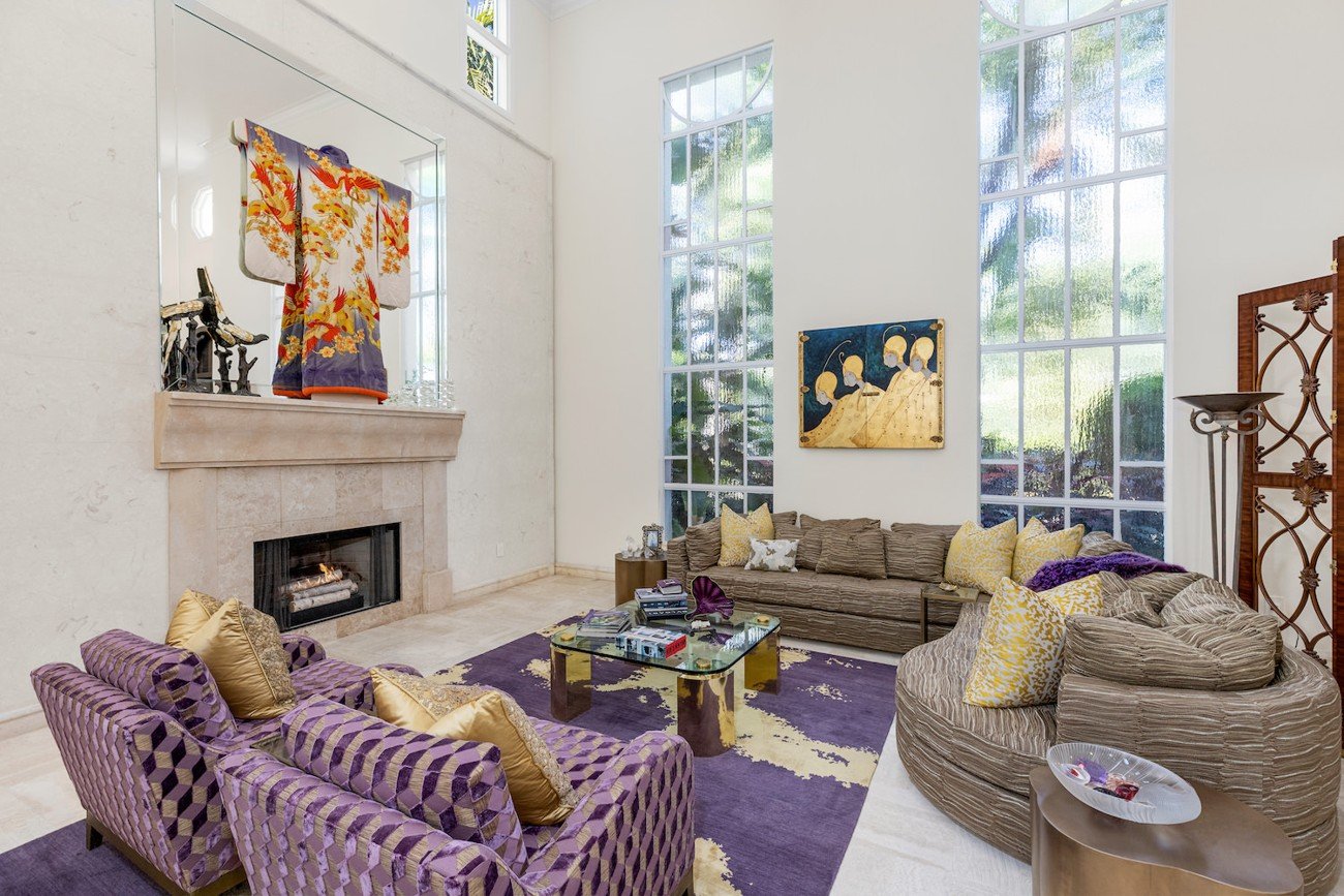 Cluttered living room with eclectic furniture, colorful artwork, and a dramatic purple rug prior to digital furniture replacement.