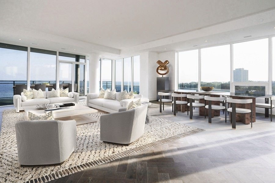 Coastal-inspired living and dining space with full-height windows, white upholstered furniture, and a chunky woven area rug facing ocean views.