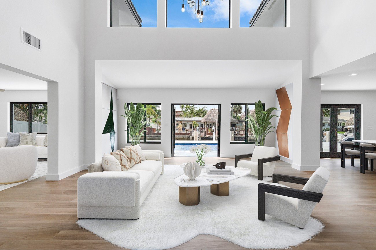 Virtually staged modern living room with white furniture, lush plants, sculptural accents, and views of the pool through tall windows.